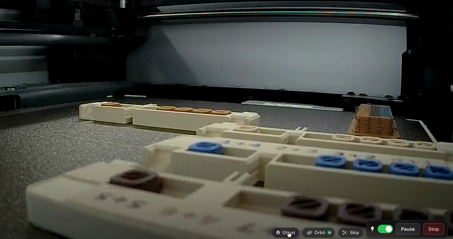 Chamber camera view of an abacus mid-print: cream printed rails on the build plate, some pockets already filled with blue and dark brown beads, an orange support tower at the back.